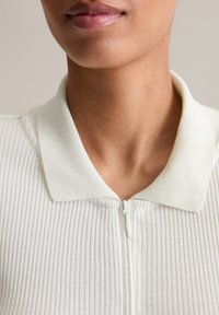 Close-up van de nek en lippen van een persoon die een witte geribbelde ritsshirt met een zachte kraag draagt.