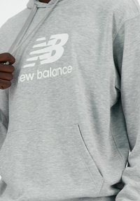 Szara bluza z kapturem z miękkiego materiału, z przednią kieszenią, kapturem ze sznurkiem i białym logo "New Balance". Gładka faktura, luźny krój.
