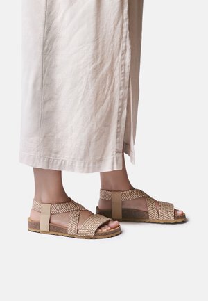 Stopy w sandałach z beżowymi plecionymi paskami i korkowymi podeszwami, zestawionych z luźnymi jasnymi spodniami z bocznym rozcięciem.