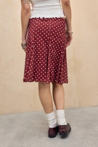 Persoană purtând o fustă roșie cu buline, top alb cu dungi, șosete albe și pantofi burgundy, stând pe un podea gri, în fața unui perete bej.