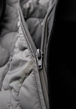 Szara pikowana tkanina o gładkim wykończeniu, z metalicznym zamkiem błyskawicznym i ciemną podszewką wewnętrzną. Zamek błyskawiczny jest funkcjonalny i elegancki.