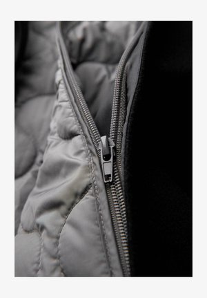 Szara pikowana tkanina o gładkim wykończeniu, z metalicznym zamkiem błyskawicznym i ciemną podszewką wewnętrzną. Zamek błyskawiczny jest funkcjonalny i elegancki.