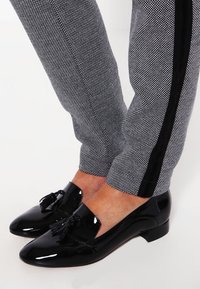 Mocassins en cuir verni noir avec un détail à gland, portés avec un pantalon à carreaux gris avec des rayures latérales noires. Talons bas.