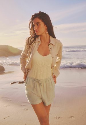 Lysfarvet linned skjorte over en tekstureret cremefarvet tanktop og lyseblå shorts med justerbar snørre, sat mod en strandbaggrund.