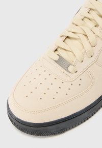 Beige ruskinds sneakers med perforeringer på tåen, tekstureret sort gummisål og et metallisk sølvmærke med "AF1."