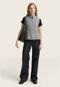 Grau gestrickter ärmelloser Pullover mit hohem Kragen, kombiniert mit einem schwarzen T-Shirt und dunklen weiten Jeans. Die Accessoires umfassen eine schwarze Handtasche.