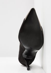 Chaussure à talon haut noire à bout pointu reposant semelle vers le haut contre une surface blanche, montrant une semelle lisse et texturée et un talon élancé.