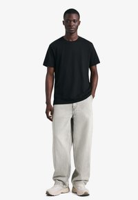 Fekete pamut rövid ujjú póló, világosszürke bő szabású farmerrel és bézs tornacipővel, fehér háttér előtt állva.