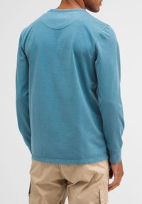 Langärmliges Hemd aus hellblauem Stoff mit rundem Ausschnitt, das eine dezente Textur und kontrastierende Nähte aufweist, getragen über beigen Cargo-Hosen.