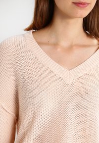 Hellrosa gestrickter Pullover mit V-Ausschnitt und lockerer Passform, mit einer strukturierten, offenen Strickstruktur und geripptem Kragen.