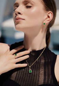 Joyería de oro con piedras verdes en corte esmeralda en un colgante, pendientes y anillo. Las piezas se combinan con una blusa negra texturizada.