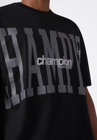 T-shirt nera con grande scritta grigia "CHAMPION" e logo bianco più piccolo "champion NEW YORK" sulla parte centrale indossata da una persona.