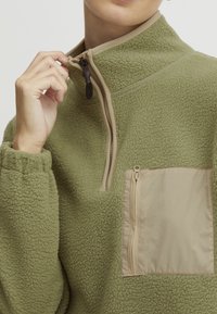 Pull-over in pile verde oliva con colletto alto, chiusura a zip e tasca con zip beige. Tessuto strutturato e maniche con polsino.