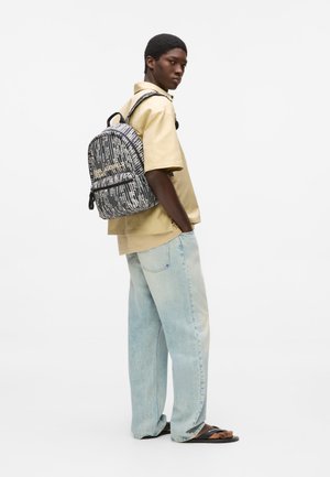 Νέος άνδρας που φοράει μπεζ πουκάμισο με κοντά μανίκια, ανοιχτό μπλε τζιν, μαύρα σανδάλια και ένα ασπρόμαυρο σακίδιο με σχέδιο.