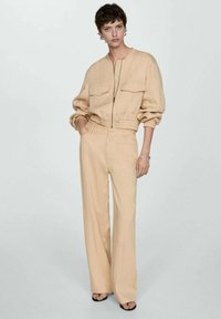 Beige, lichtgewicht jumpsuit met een rits aan de voorkant, twee borstzakken en een elastische taille; heeft een wijde broekspijp en een comfortabele pasvorm.