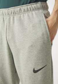Sive trenirke iz mehkega materiala, z elastičnim pasom in črnim Nike logotipom na levi strani. Gladka tekstura in sproščen kroj.