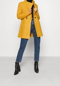 Abrigo amarillo texturizado con cuello, dos botones, bolsillos delanteros, combinado con jeans azules y botas negras de tacón.