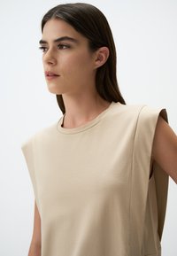Beżowy top bez rękawów, wykonany z gładkiego materiału, z okrągłym dekoltem i delikatnymi akcentami na ramionach. Prosty, minimalistyczny design.
