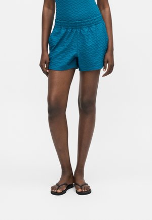 Femme portant un short texturé bleu sarcelle et un haut assorti, debout pieds nus avec des tongs noires sur un fond uni.