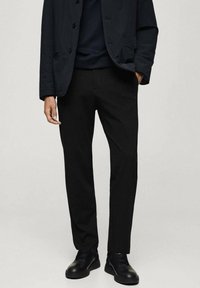 Man in zwarte op maat gemaakte broek, zwarte jas, zwart sweater en zwarte veterschoenen, staande met één hand in de zak.