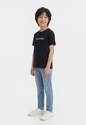 Boy wearing a black Calvin Klein t-shirt, light blue jeans, and white sneakers, standing and smiling, looking to the side.