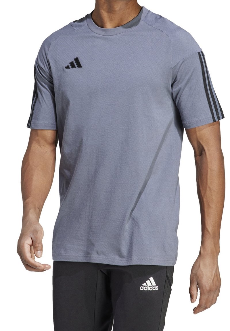Homme portant un t-shirt à manches courtes gris Adidas avec des bandes noires sur les manches et un pantalon noir Adidas avec un logo sur la cuisse gauche.