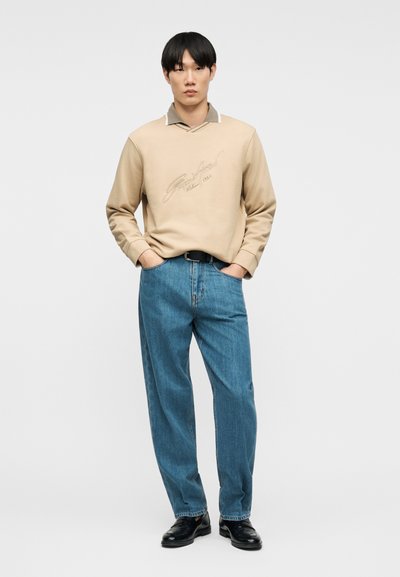 Νέος άνδρας που στέκεται με τα χέρια στις τσέπες, φορώντας μπεζ πουκάμισο με μακριά μανίκια και γιακά, μπλε τζιν, μαύρη ζώνη και μαύρα παπούτσια σε λευκό φόντο.