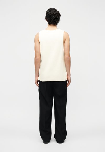 Άνδρας με σγουρά μαλλιά που φοράει λευκό αμάνικο πάνω μέρος και φαρδιά μαύρα παντελόνια, στέκεται με την πλάτη προς τα εμπρός σε λευκό απλό φόντο.