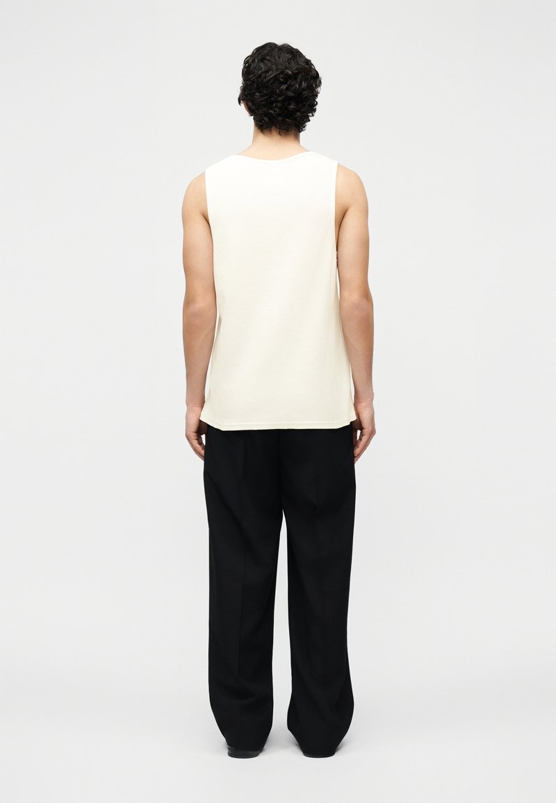 Uomo con capelli ricci che indossa un top bianco senza maniche e pantaloni neri larghi, in piedi di spalle su uno sfondo bianco neutro.