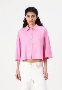 Rosa, kortermet, skjorte med krage og knapper nedover fronten; løs passform og litt cropped kant. Myk tekstur med en glatt overflate.
