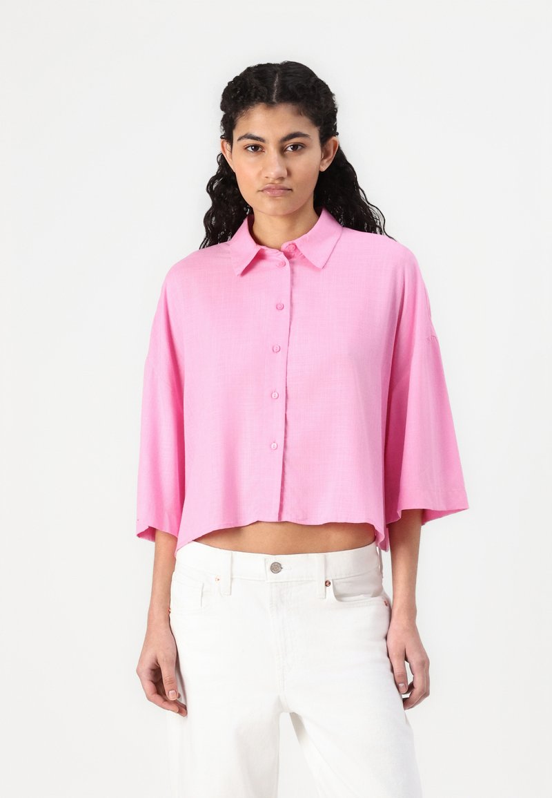 Rosa, kortermet, skjorte med krage og knapper nedover fronten; løs passform og litt cropped kant. Myk tekstur med en glatt overflate.