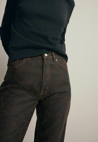 Jeans marrón con un patrón, fabricados con un tejido texturizado, que cuentan con detalles cosidos y un cierre frontal de botón, combinados con una blusa negra ajustada de manga larga.