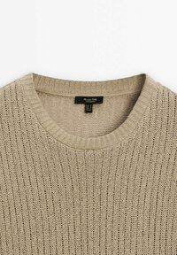 Beige neulepaita, jossa on ribbitekstuuri, pyöreä pääntie ja merkki, esittelee pystysuoria viivoja koko kankaan läpi.