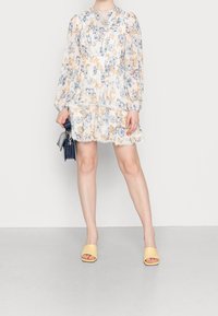 Blommig miniklänning i lätt tyg, långa puffärmar, volangkant och spetsdetaljer i pastellfärger; tillsammans med gula slip-in sandaler.