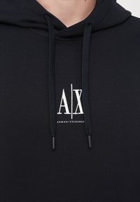 Marineblaues Kapuzensweatshirt mit schwarzen Kordeln, das ein weißes Logo "A|X" und den Text "ARMANI EXCHANGE" auf der Brust zeigt. Glatte Stoffoberfläche.