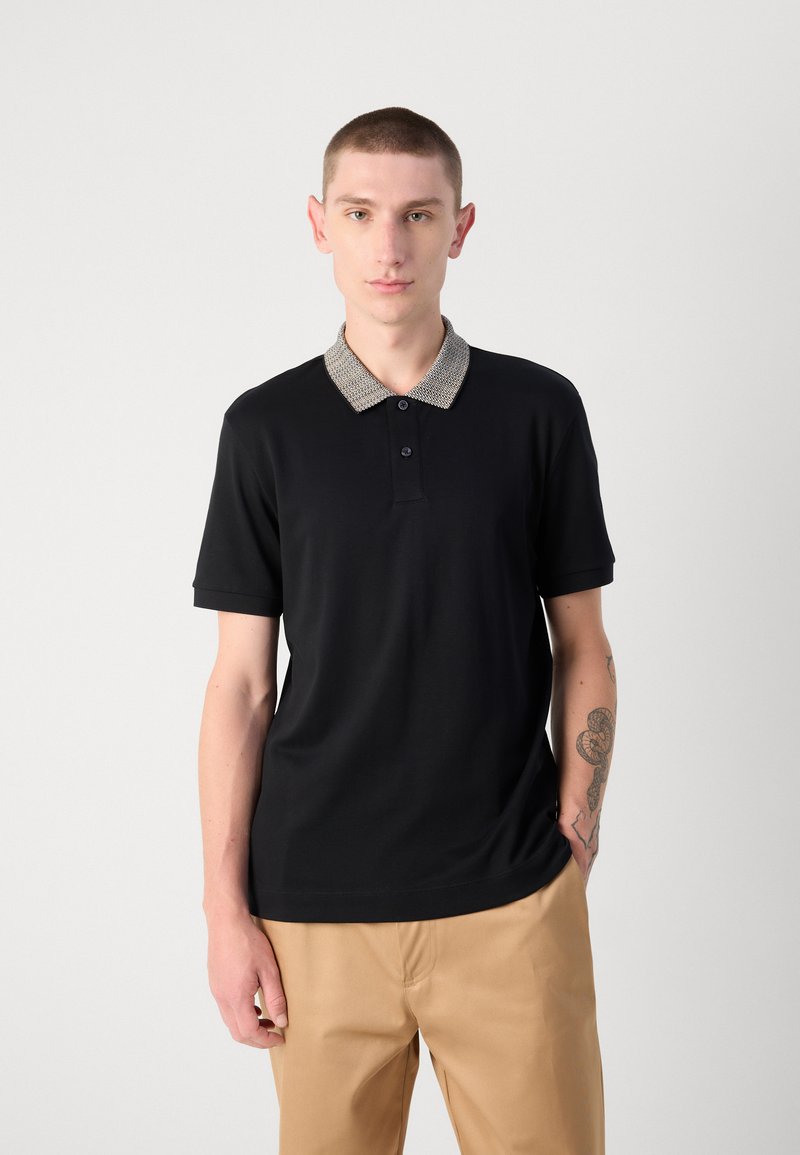 Schwarzes Poloshirt mit einem gemusterten Kragen, zwei Knöpfen am Hals und kurzen Ärmeln, kombiniert mit hellbeigen Hosen.
