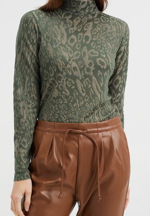 Zielony golf w zwierzęcy wzór noszony z brązowymi spodniami z wysokim stanem z sztucznej skóry, ozdobionymi przednim ściągaczem i zakładkami.