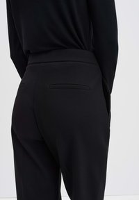 Zwarte broek van soepel materiaal met een getailleerde pasvorm, voorzien van twee achterzakken en een vereenvoudigd ontwerp van de tailleband.