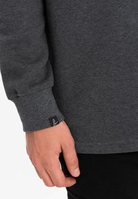 Grijze longsleeve shirt van een gestructureerde stof, met gestikte manchetten en een klein zwart labeldetail op de mouw. Hand gedeeltelijk zichtbaar.