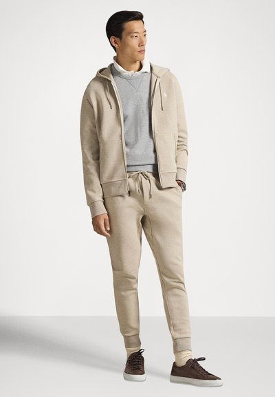 Μπεζ φούτερ με φερμουάρ και κορδόνια, γκρι φούτερ από κάτω και τζόγκερ στο ίδιο μπεζ χρώμα. Καφέ παπούτσια ολοκληρώνουν την εμφάνιση.