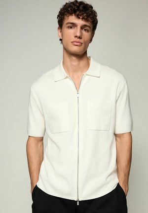 Krátkozrý krátky krémový zip-up košeľa vyrobená z jemného materiálu, s dvoma prednými vreckami a plochým golierom. Hladká textúra s uvoľneným strihom.