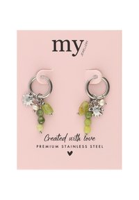 Orecchini in acciaio inossidabile con gemme verdi, piccoli ciondoli a forma di sole in argento e diverse forme di perline. Presentati su un cartoncino rosa con branding.