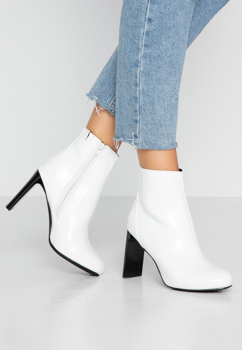 Weiße Lackleder-Ankle Boots mit hohem Blockabsatz, ausgestattet mit einem seitlichen Reißverschluss und einer glatten Oberfläche. Kombiniert mit Jeans mit fransigem Saum.