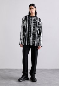 Čierno-biela košeľa s dlhým rukávom s výrazným grafickým vzorom, skombinovaná s čiernymi nohavicami a robustnými čiernymi teniskami.