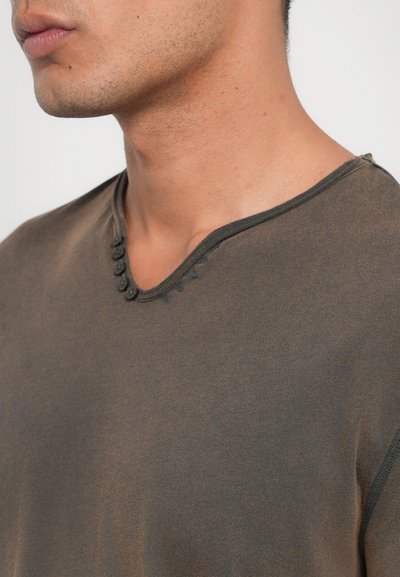 Brown, short-sleeve shirt with a v-neckline. Features decorative buttons along the neckline and a soft, textured fabric.