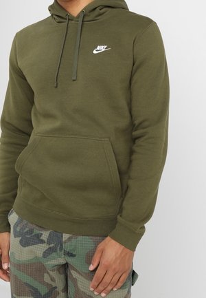 Homme portant un sweat à capuche Nike vert olive avec poche avant et pantalon cargo camouflage, debout sur un fond blanc.