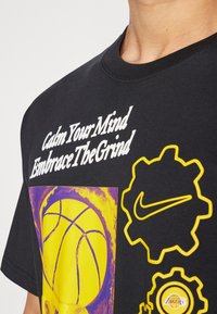 Černé tričko s nápisem "Uklidni svou mysl, přijmi námahu", grafickým prvkem žlutého basketbalového míče a logem Nike uvnitř tvarů ozubeného kola. Materiál: bavlna.