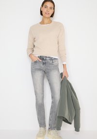 Pull beige, jeans gris délavés, tenant une veste bomber verte. Chaussures claires, style décontracté, design simple, avec une texture douce.