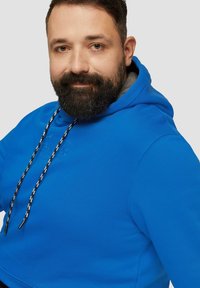 Niebieska bluza z kapturem z kieszenią kangurką. Czarno-białe sznurki i wytłaczany detal logo na klatce piersiowej. Miękka, gładka tekstura.