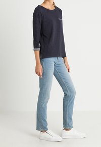 Navy polka dot top with three-quarter sleeves and a pocket, paired with light blue jeans and white sneakers. Simple, casual design.